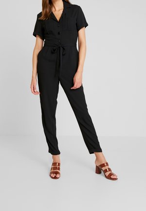 Kvinde iført en sort kortærmet button-up overall med bundet talje og brune hælede sandaler, stående mod en ensfarvet grå baggrund.