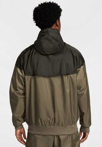 Oliwkowa kurtka z kapturem, z ciemniejszą górną częścią, elastycznymi mankietami i dołem. Materiał wydaje się gładki, o matowym wykończeniu.