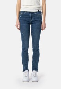 Mršave traperice u tamnoplavoj denim boji s istrošenim detaljima i klasičnim dizajnom s pet džepova, nosi se s bijelim tenisicama.