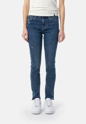 Jean slim en denim bleu foncé avec des accents déchirés et un design classique à cinq poches, portés avec des baskets blanches.