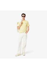 Geel katoenen t-shirt met een zak, gedragen met witte wijde broek en beige sneakers. Model houdt een witte jas vast en draagt zwarte zonnebril.