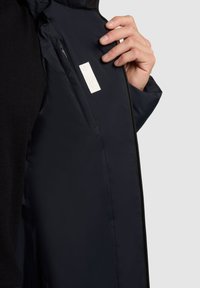 Tumed sinine jakk sileda, mati viimistlusega, mis sisaldab lukustatavat sise-taskut ja väikest valget silti. Käsi on paigutatud lähedale avamisele.