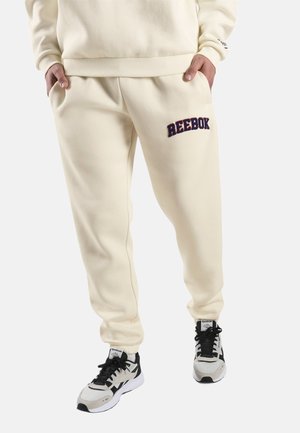 Persoon draagt roomkleurige Reebok joggingbroek met logo op de dij, gecombineerd met witte en zwarte Reebok sneakers, staand met handen in de zakken.