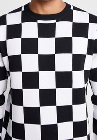 Homme portant un pull à manches longues noir et blanc à carreaux avec un col rond.