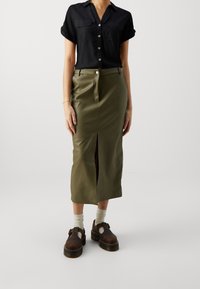 Black button-up shirt with rolled sleeves. Olive green leather midi skirt with front slit. Brown shoes with straps and white socks.