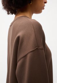 Brun bomulds sweatshirt med en afslappet silhuet, sænkede skuldre og synlige stikninger på en neutral baggrund.