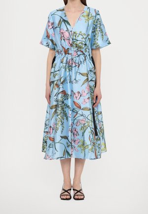 Femme portant une robe midi bleu clair avec imprimé floral rose et vert, manches courtes, col en V, taille froncée et sandales noires à lanières.