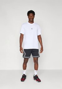 Mann som står iført T-skjorte med hvit Jordan-logo, svarte basketballshorts med hvit kant, hvite Nike-strømper og svarte-røde sneakers.