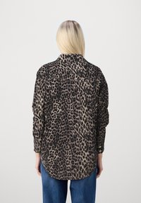 Blonde Person in einem langärmeligen Leopardenmusterhemd und blauen Jeans, mit dem Rücken zur Kamera stehend vor einem schlichten Hintergrund.