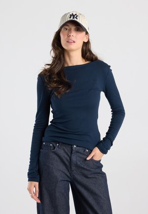 Vrouw met lang bruin haar draagt een marineblauw shirt met lange mouwen, krijtstreepbroek en een beige baseballpet met een donker logo, staand met één hand in de zak.