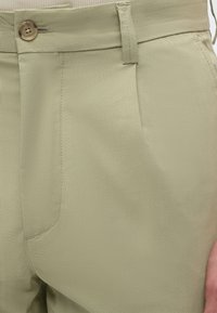 Gros plan sur la taille d’un pantalon beige avec fermeture à bouton, passants de ceinture, poche latérale et détails visibles des coutures.