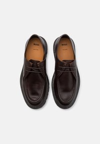 Chaussures en cuir marron foncé à finition lisse, forme arrondie, détails de couture subtils et semelles en caoutchouc. Marque visible à l'intérieur.