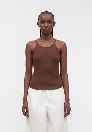 Donna con capelli intrecciati che indossa una canotta marrone e pantaloni bianchi larghi, in piedi davanti a uno sfondo grigio chiaro uniforme.