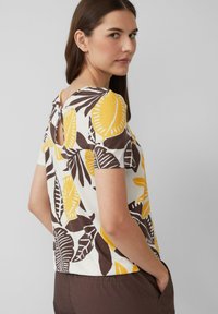 T-shirt blanc à manches courtes avec des motifs de feuilles jaunes et brunes, col rond et un détail en forme de trou de serrure à l'arrière. Tissu en coton doux.