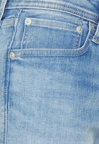 Ljusblå denim med mjuk textur, har en framficka, metallnitar och kontrasterande sömmar längs kanterna.