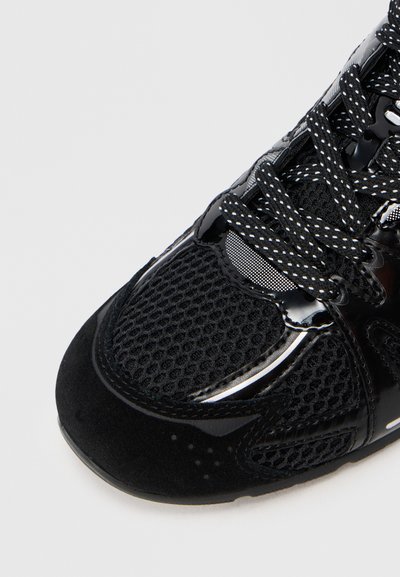 Zapatilla deportiva negra con detalles de malla y piel sintética, con cordones negros y blancos con manchas atados en forma de lazo.