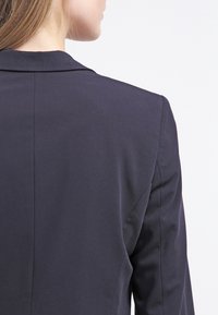 Tmavě navy sako s přiléhavým střihem, se šálovým límcem a hladkým, texturovaným materiálem, zobrazené zezadu. Jednoduché a elegantní.