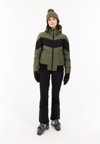 Groene en zwarte gewatteerde jas met een chevronontwerp, zwarte handschoenen, zwarte broek en grijze skiboots. Vergezeld van een gebreide olijfgroene muts.