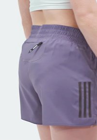 Lila Sportshorts aus leichtem Material, mit elastischem Bund, einer Reißverschlusstasche auf der Rückseite und drei schwarzen Streifen an der Seite.