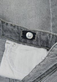 Pilka džinsinė medžiaga su matoma juoda juosmens mygtuku. Yra maža vidinė balta kišenė ir siuvinėjimas palei kraštus.