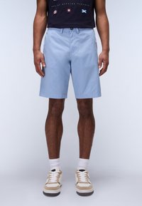 Shorts bleu clair en tissu lisse, coupe ajustée, avec poches latérales, associés à des baskets beiges et des chaussettes blanches.