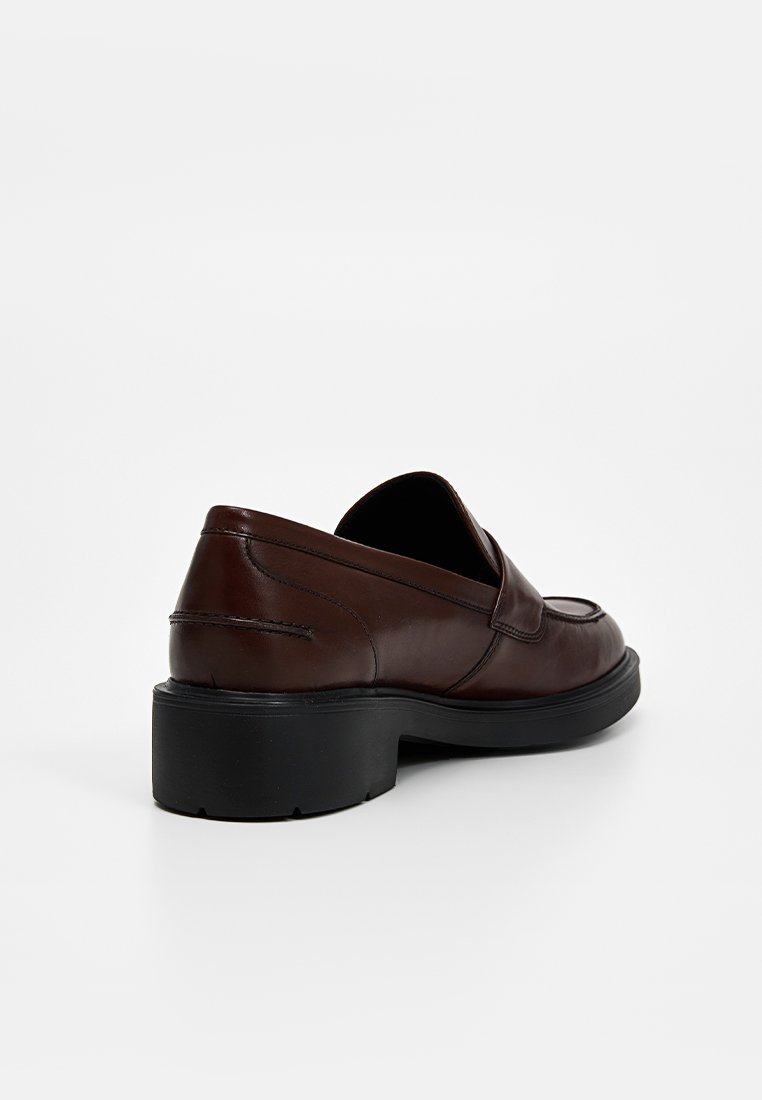 Chaussure slip-on en cuir marron avec une semelle noire texturée basse et des coutures courbes, présentant un design minimaliste sans matériel visible.