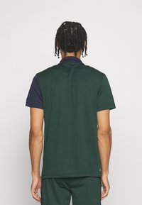 Man med flätat hår som bär en mörkgrön och marinblå kortärmad polo och matchande byxor, stående vänd bort från kameran mot en enfärgad bakgrund.