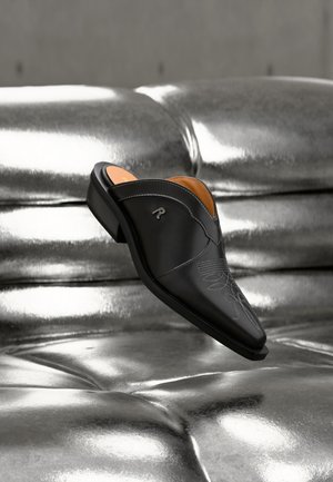 Μαύρο δερμάτινο παπούτσι slip-on με διακοσμητικό ράψιμο, τοποθετημένο πάνω σε γυαλιστερή ασημένια επιφάνεια με καπιτονέ μοτίβο, γωνιακά τοποθετημένο για να φαίνονται οι λεπτομέρειες από το πλάι και από πάνω.