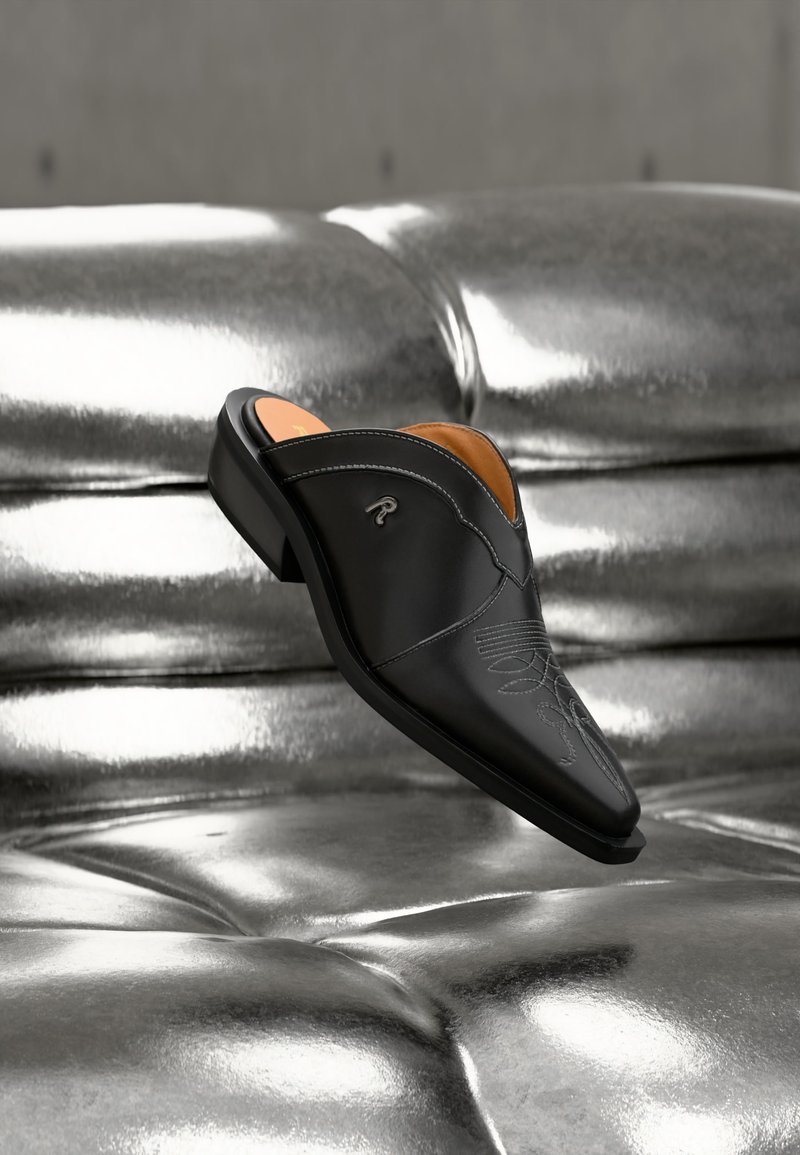 Μαύρο δερμάτινο παπούτσι slip-on με διακοσμητικό ράψιμο, τοποθετημένο πάνω σε γυαλιστερή ασημένια επιφάνεια με καπιτονέ μοτίβο, γωνιακά τοποθετημένο για να φαίνονται οι λεπτομέρειες από το πλάι και από πάνω.