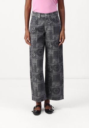 Persoon die zwarte wijduitlopende jeans draagt met lichtgrijs patchworkpatroon, roze top en zwarte Mary Jane-schoenen met gespen op witte achtergrond.