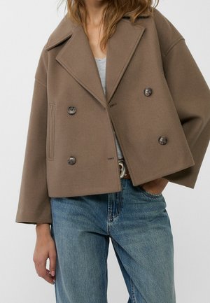 Vrouw draagt een cropped, bruin, dubbelgeknoopt wollen jas over een grijze top en blauwe spijkerbroek met een bruine leren riem.