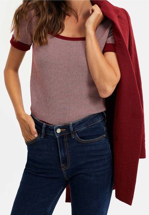 Femme portant un haut à manches courtes à motifs rouges et blancs, un jean bleu foncé, tenant une veste rouge sur l'épaule.