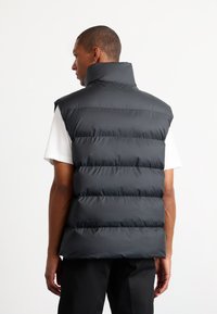 Sort polstret vest med høj krave, ærmeløs design og vandret syninger, båret over en hvid skjorte mod en ensfarvet baggrund.
