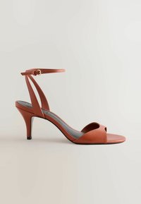 Sandale à talon haut en cuir marron à bout ouvert avec bride à la cheville et petite boucle dorée sur fond gris clair.