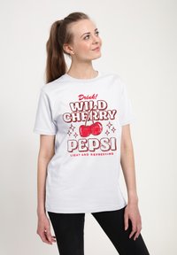 B�ílá bavlněná tričko s výrazným červeným nápisem "WILD CHERRY PEPSI" s dvěma třešněmi a hvězdami; nošeno s černými kalhotami.