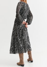 Robe noire à motif de fougère blanche, manches longues en voile transparent et taille cintrée, associée à des bottes mi-mollet marron clair à talon bas.
