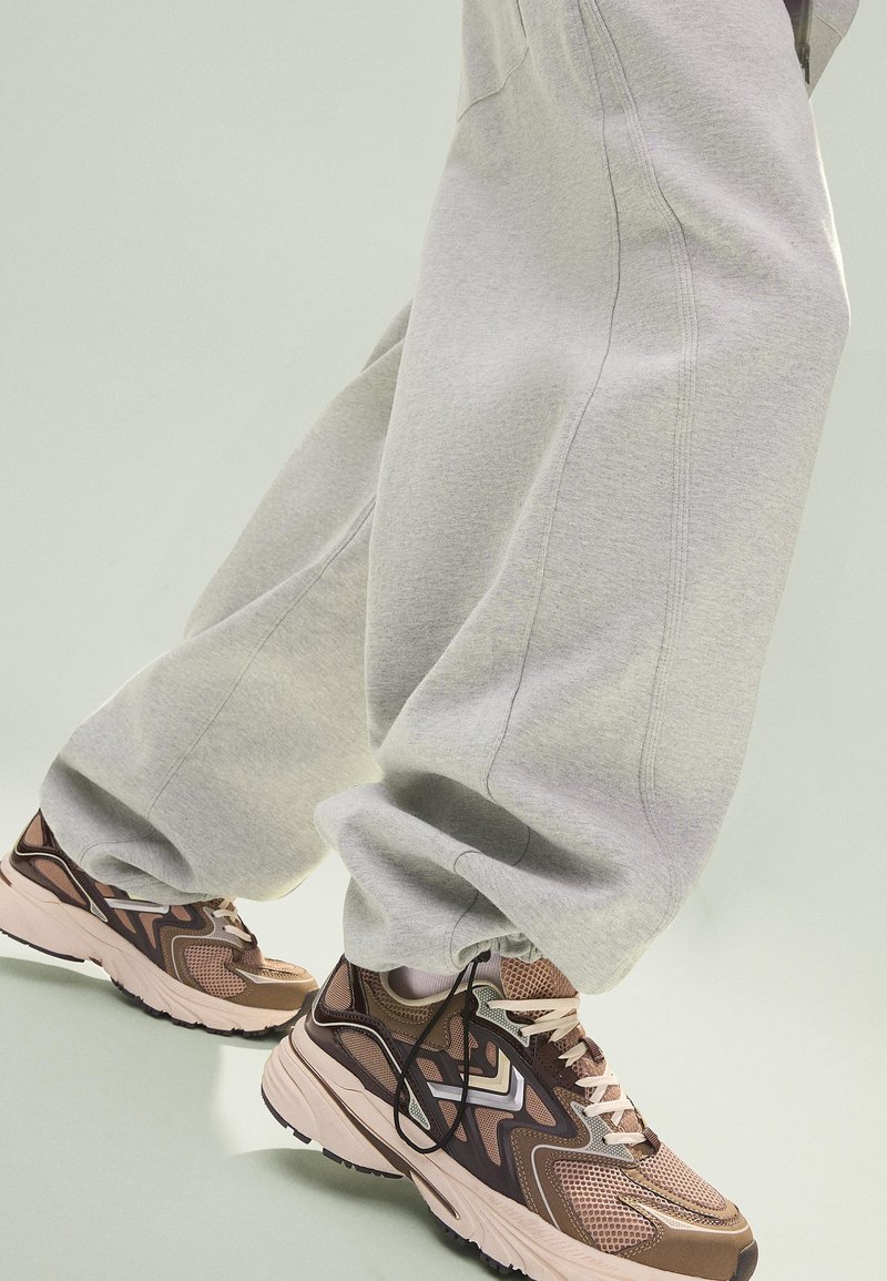Piedi con sneakers in mesh marrone e beige con accenti neri e lacci bianchi, abbinati a pantaloni della tuta larghi di colore grigio chiaro.