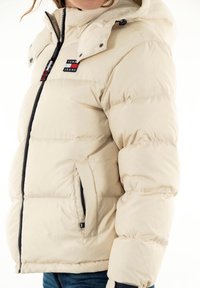 Beige pufferjacka med quiltad struktur, avtagbar huva, framdragkedja och logotypdetalj. Har sidodragkedjefickor och elastiska ärmslut.