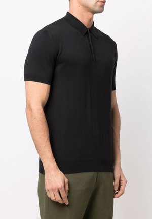 Uomo che indossa una polo nera aderente a maniche corte e pantaloni verde oliva, in piedi davanti a uno sfondo bianco uniforme.