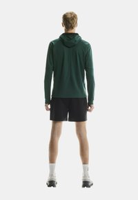 Mann, der mit dem Rücken zur Kamera steht, trägt eine dunkelgrüne Kapuzenjacke, schwarze Shorts, weiße Socken und weiße Laufschuhe vor einem weißen Hintergrund.