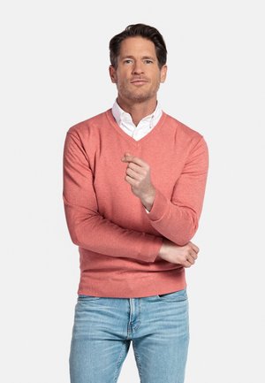 Man met bruin haar die een koraalkleurige V-hals trui draagt over een wit overhemd, lichtblauwe jeans, met één hand bij zijn borst, staand tegen een witte achtergrond.