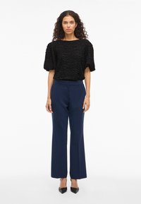 Melnas teksturētas blūzes ar īsām puffētām piedurknēm komplekts ar piegrieztām tumši zila biksēm un melnām norādītām papēžkurpēm, stāvot pret baltu fona.