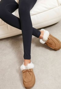 Persona vestida con leggins negros sentada en un sofá blanco, con los pies cruzados, usando zapatillas mocasín color beige forradas con piel blanca.