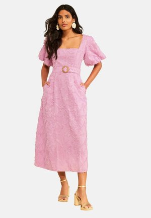 Femeie care poartă o rochie midi texturată lavandă cu mâneci bufante, decolteu pătrat, curea cu cataramă circulară și sandale bej cu toc bloc.