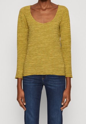Woman wearing a fitted mustard yellow ribbed long-sleeve top with a scoop neckline and dark blue jeans, standing against a plain background.
