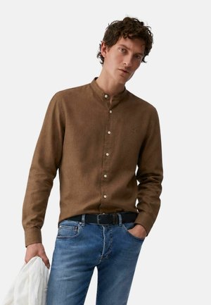 Uomo che indossa una camicia marrone con bottoni e jeans blu con cintura nera, che tiene una giacca bianca in una mano, su uno sfondo semplice.