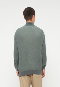 Pulover tricotat verde, cu guler înalt, croială lejeră și mâneci lungi, având o textură solidă și un tiv contrastant.
