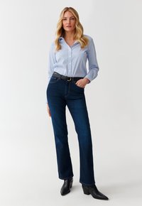 Chemise boutonnée bleu clair à rayures blanches, associée à un jean bleu foncé et des bottines noires, présentant une coupe ajustée et une ceinture classique.