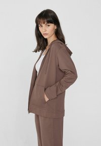 Sudadera con cremallera en color marrón, con capucha ajustable, bolsillos laterales y mangas largas. La tela parece suave y lisa. Se lleva sobre una camiseta sin mangas blanca.
