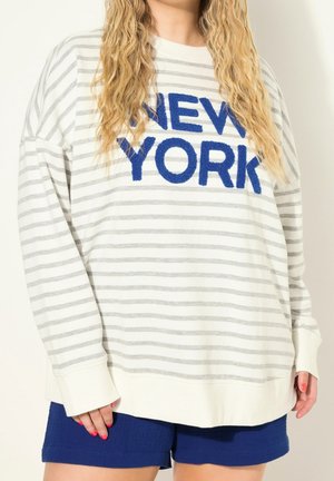 Person iført en hvid og gråstribet langærmet skjorte med "NEW YORK" i blå bogstaver og blå shorts, med synlig rød neglelak.
